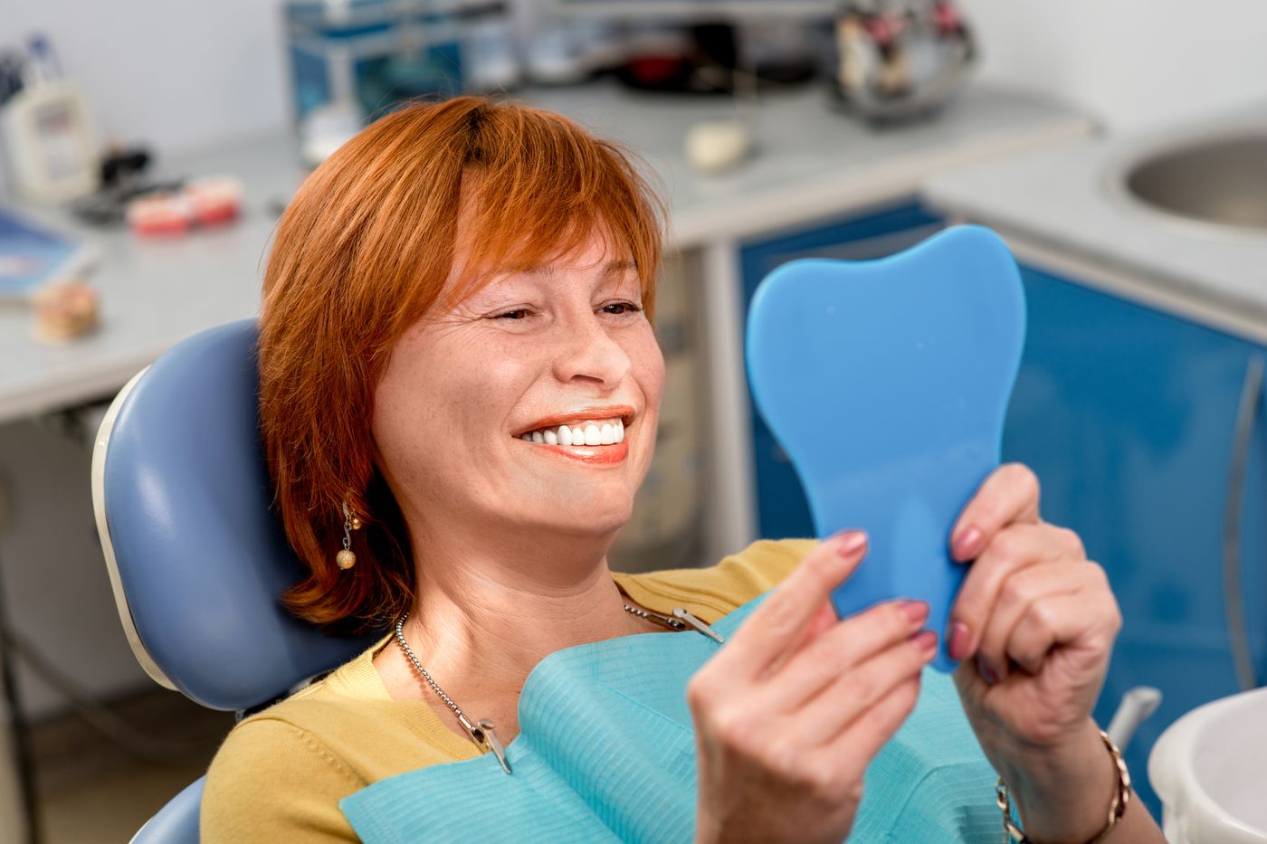 Smiling woman in dental chair holding blue tooth-shaped mirror for self-examination