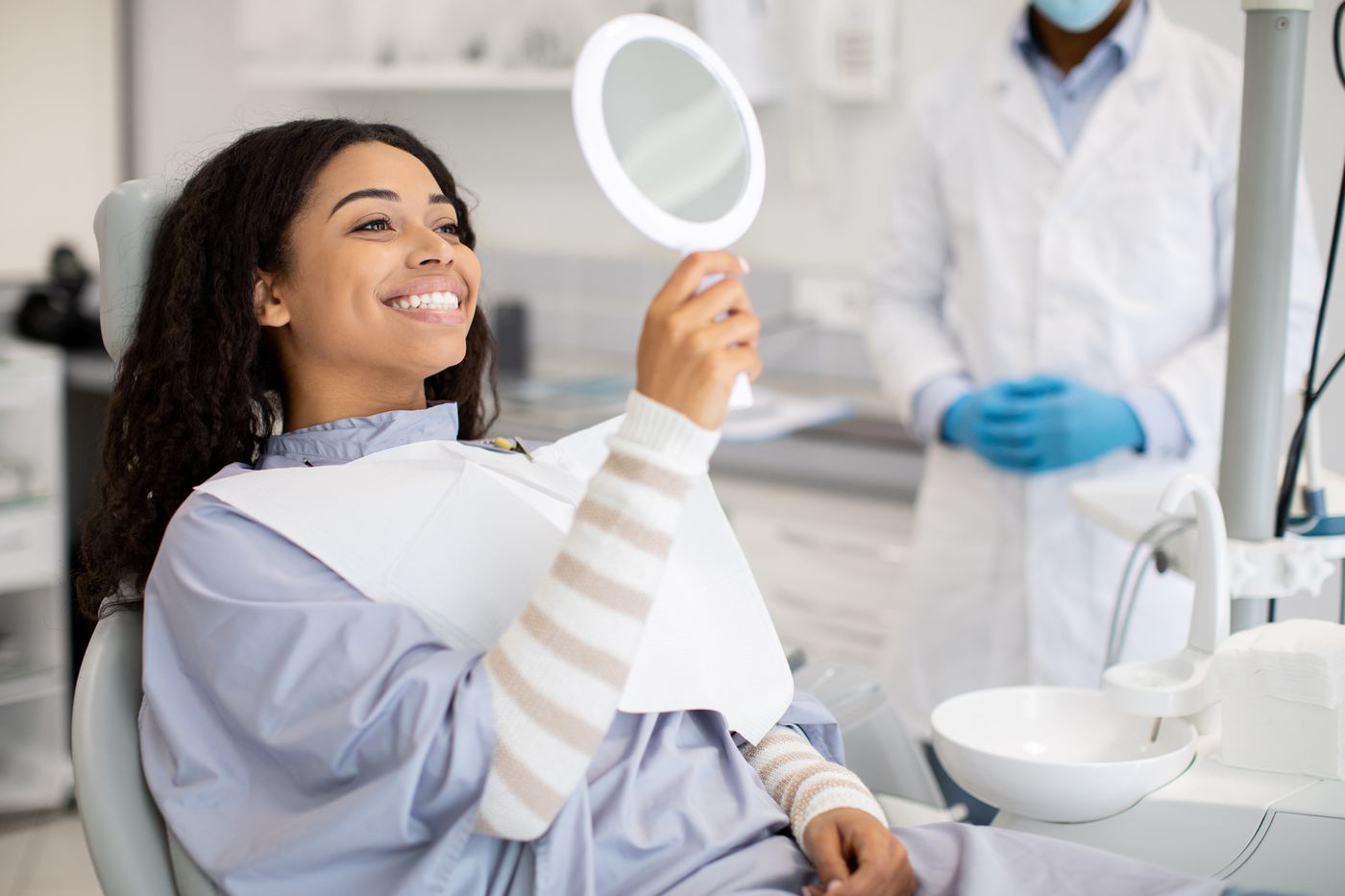 Smiling patient in dental chair holding round mirror with dentist nearby